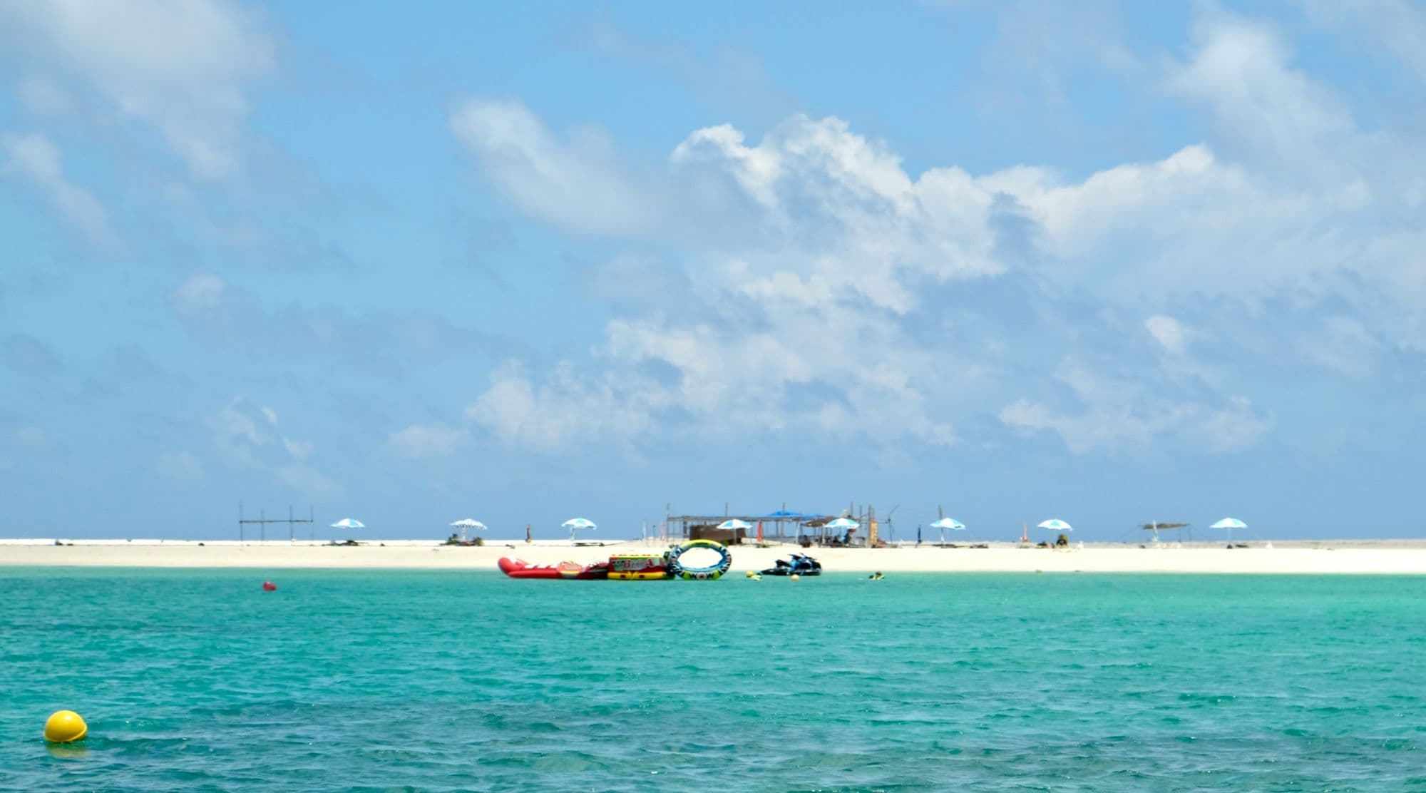 White sandbar with beach umbrellas and boats anchored in turquoise shallows