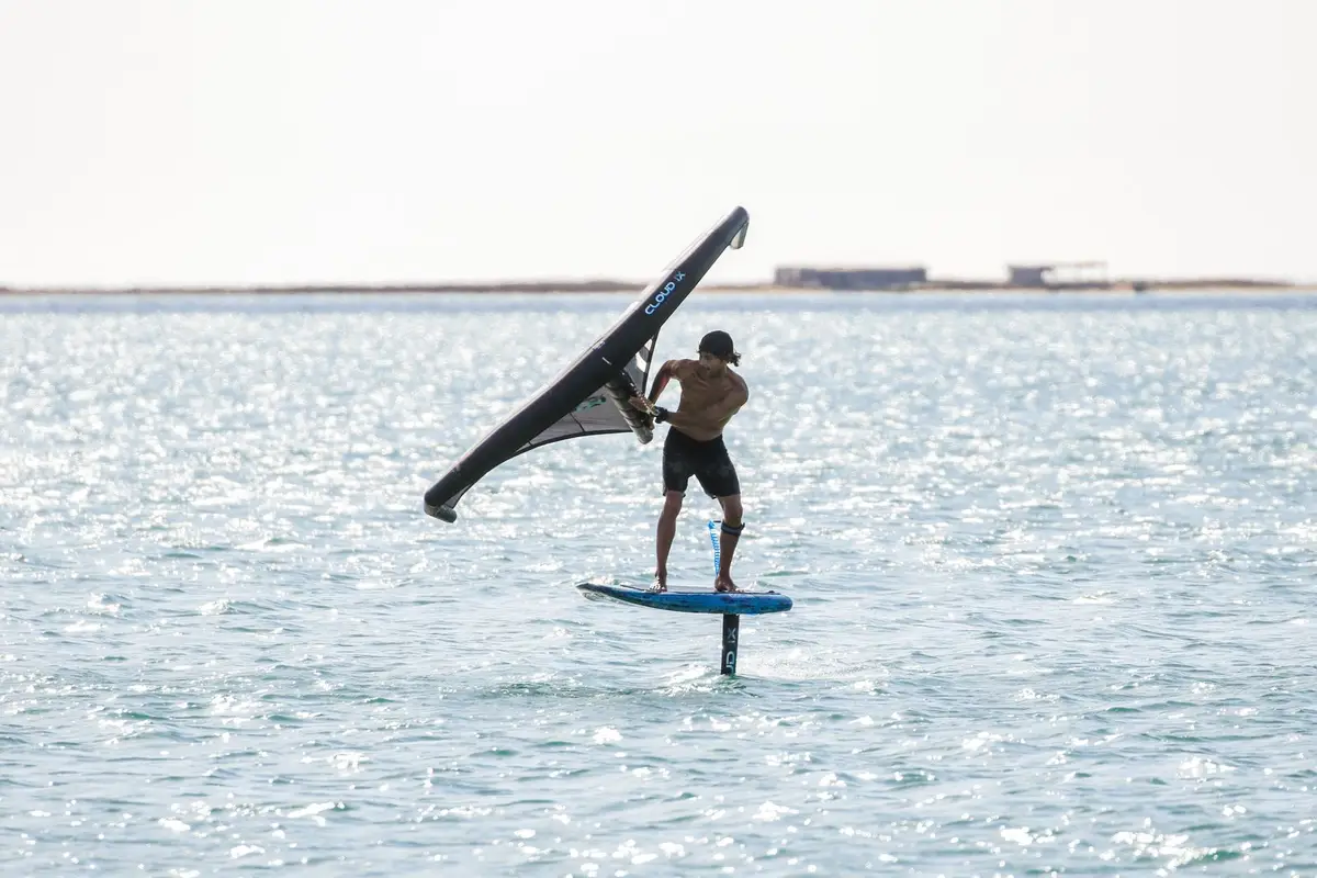 A wing foiler holding the wing overhead while riding a hydrofoil board on calm, sunlit water