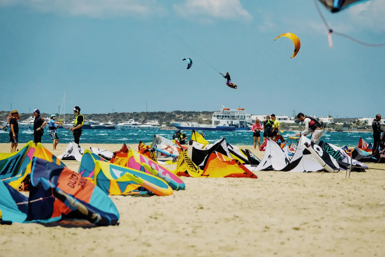 Kites laid out on a busy beach while riders head out on the water