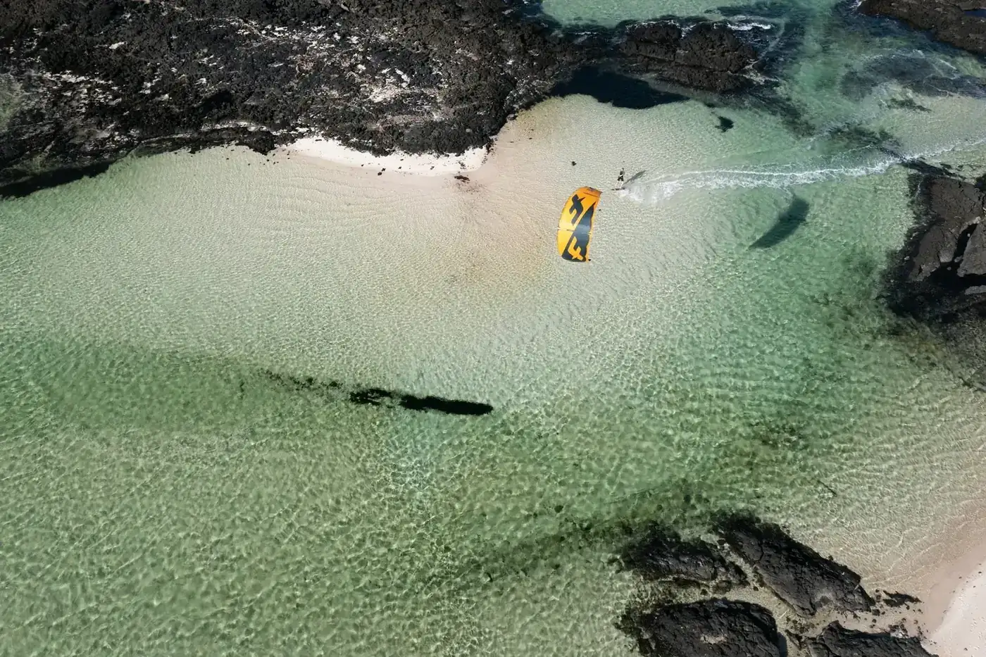 A kitesurfer over clear shallow water between dark lava rocks