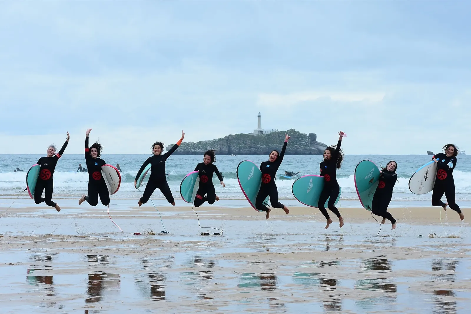 A group of Escuela Cántabra de Surf students jumping with their boards on Somo beach, a lighthouse island behind them