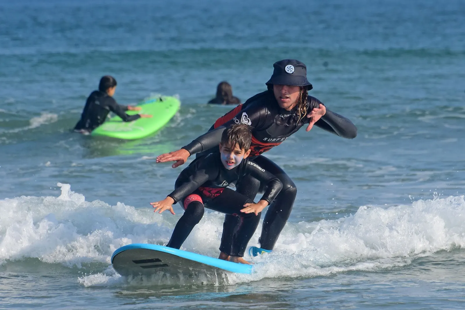 An Escuela Cántabra de Surf instructor riding a small wave behind a young student