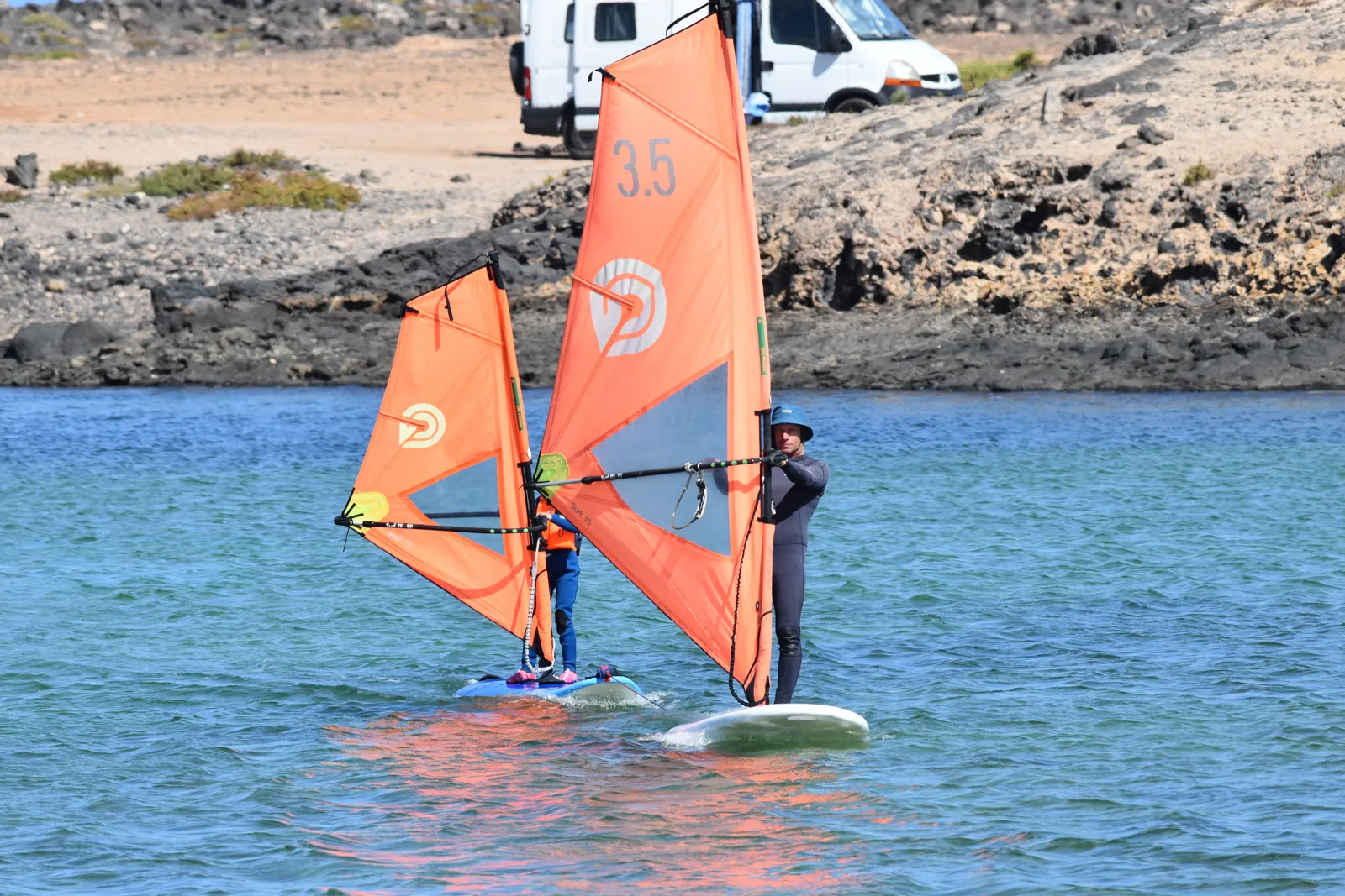 A Gecko instructor guiding a beginner on a windsurf board in the lagoon
