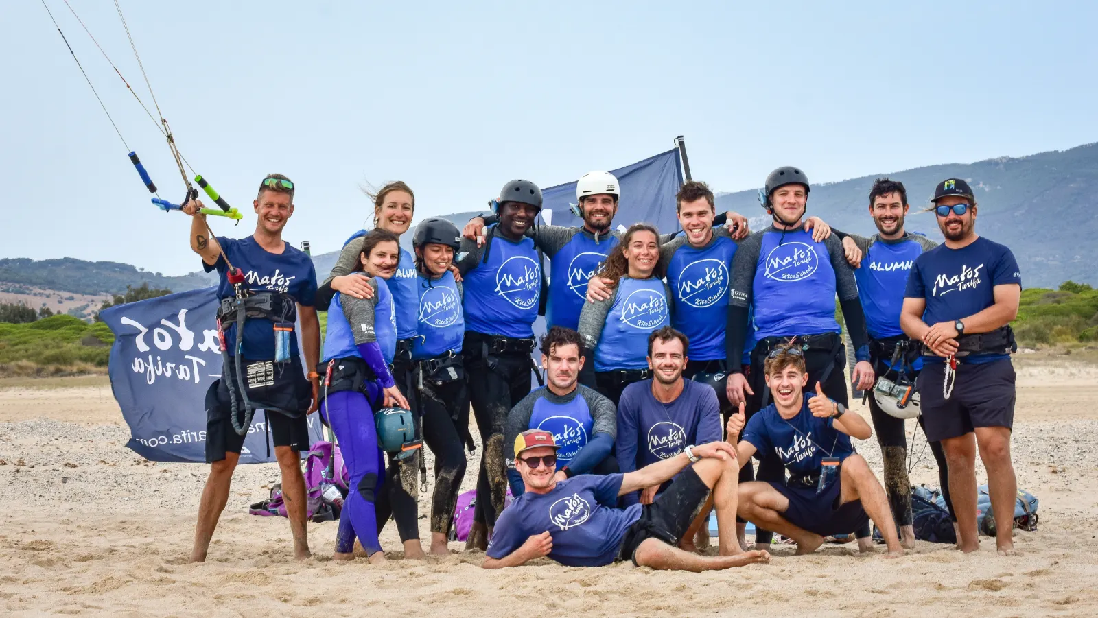 The Matos Tarifa team and students in blue lycras posing on the beach next to the school's flag
