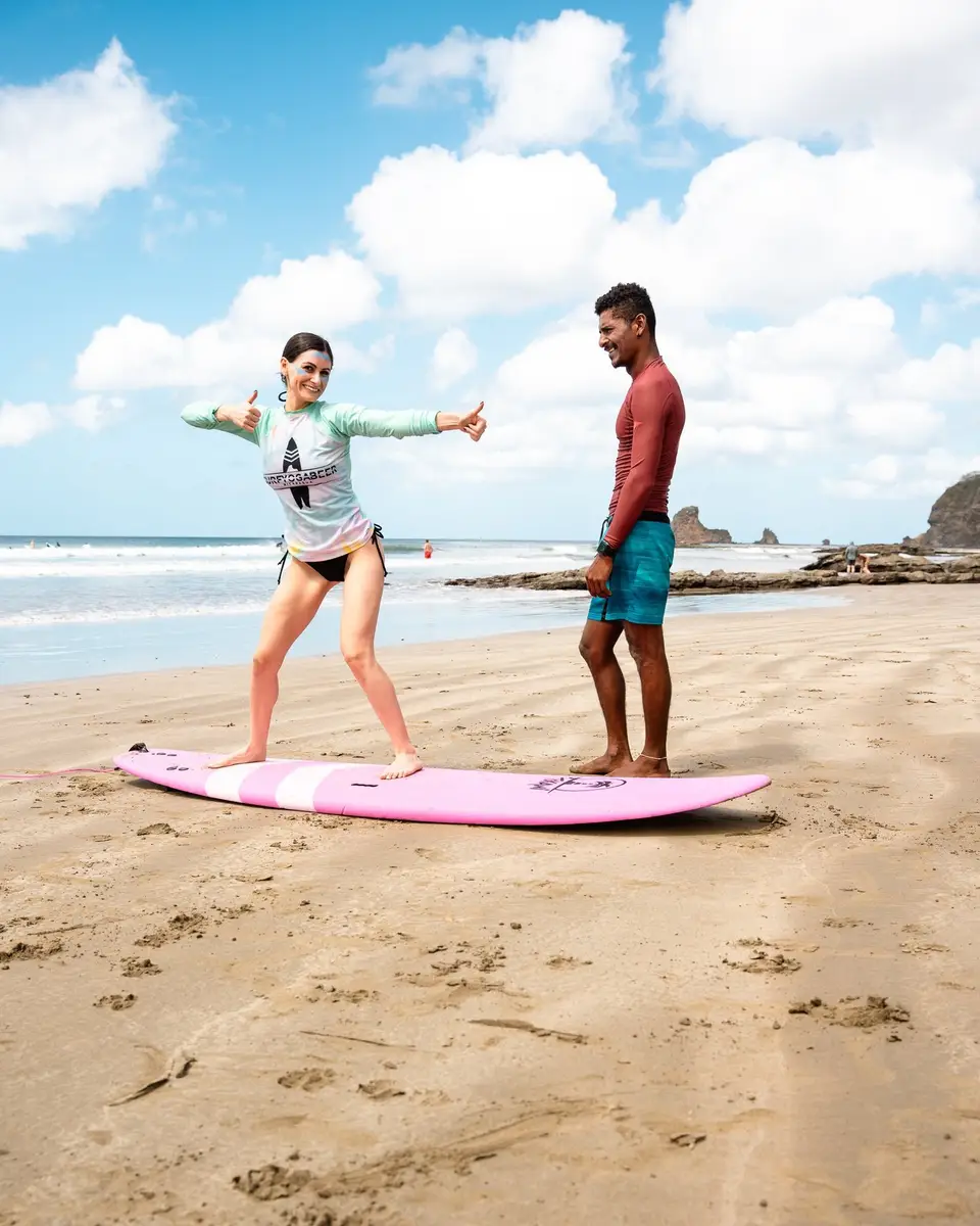 A Beach Break Surf School coach watching a student stand on a pink surfboard on the sand at Playa Maderas