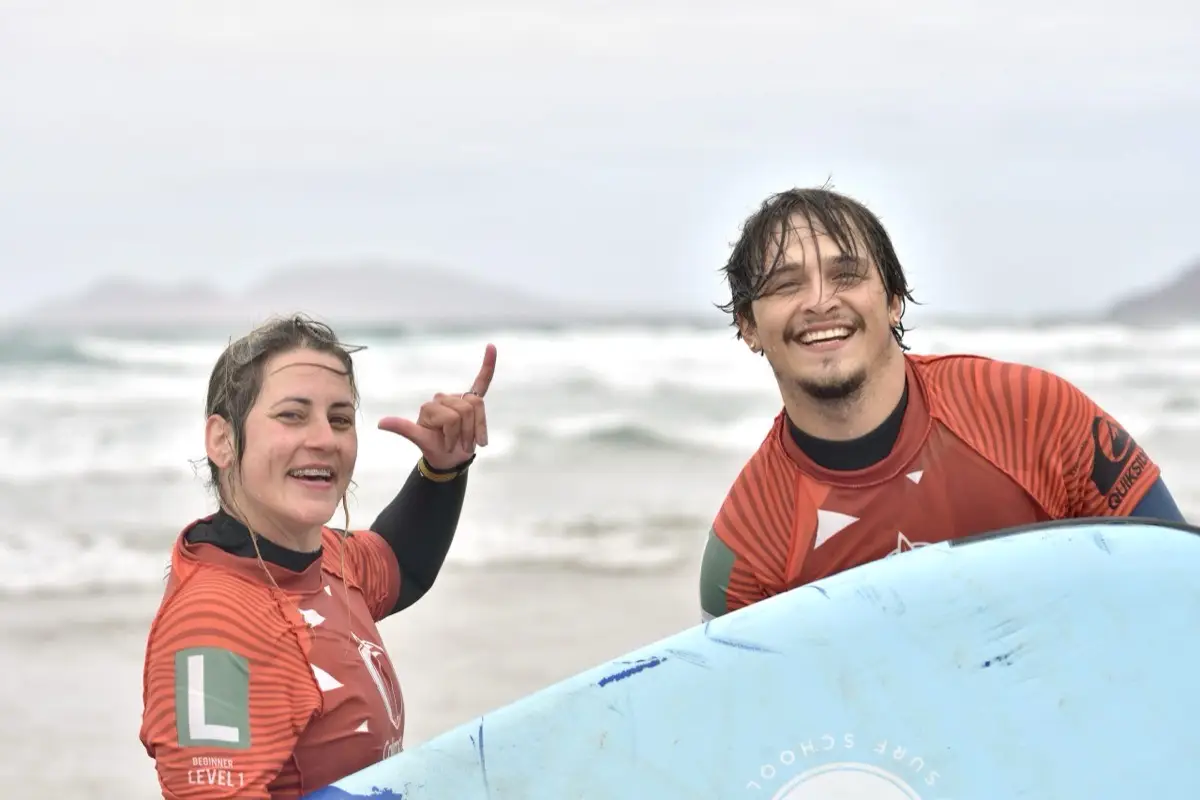 Two Calima students in school lycras smiling with a surfboard on Famara beach