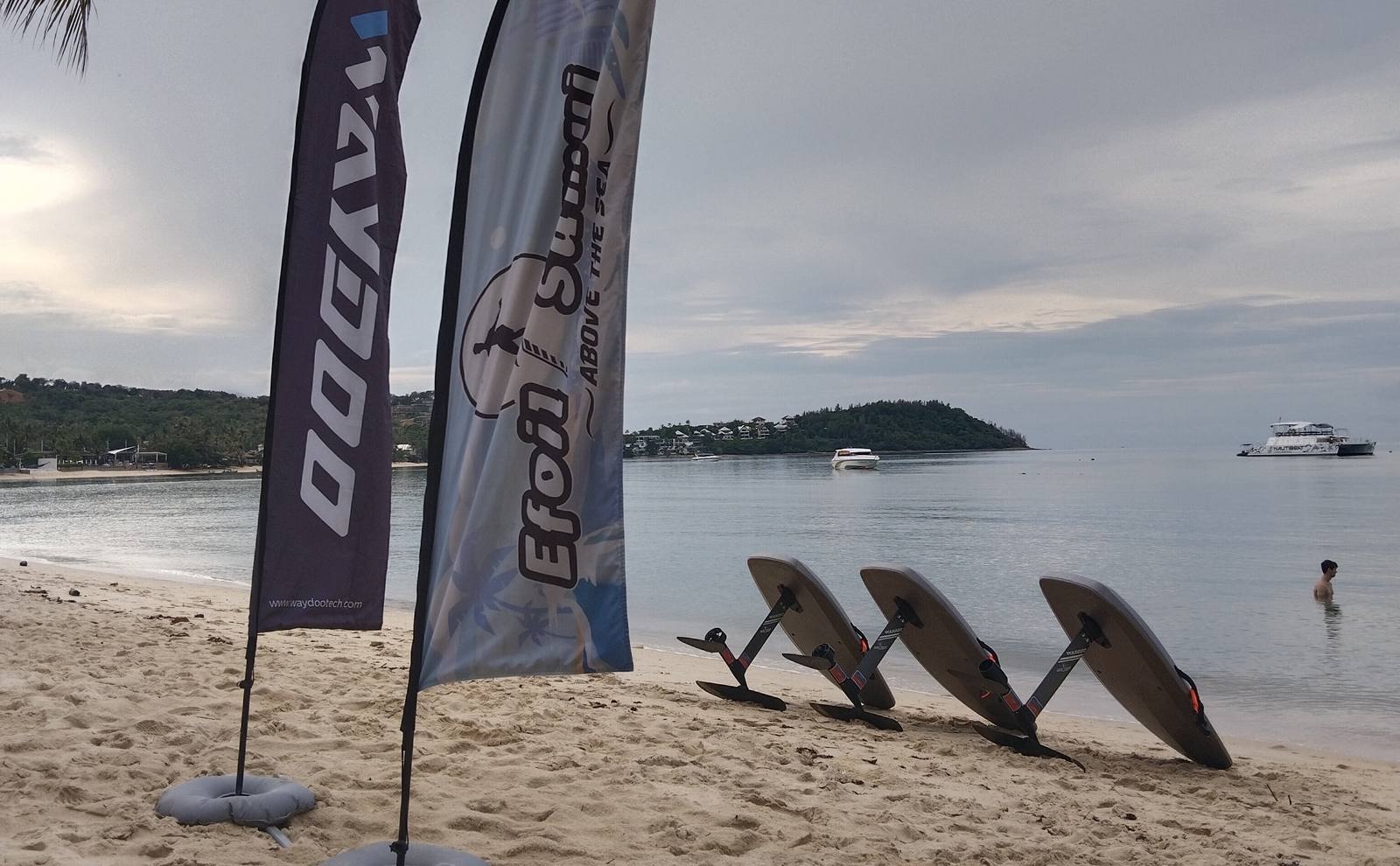 WayDoo and Efoil Samui flags with three e-foil boards on the sand, the bay behind