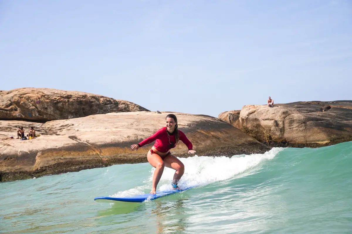 A student in a red school rash guard standing up on a blue board on a small wave, rocks behind
