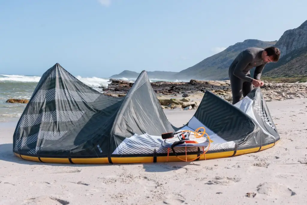 A kite and its bar laid out on the sand, a rider packing down in front of the mountains