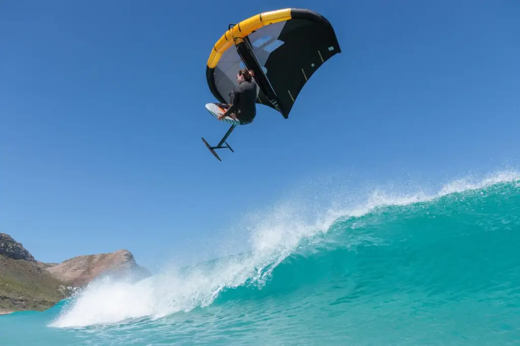 A wing foiler flying above a turquoise wave with a yellow and black wing