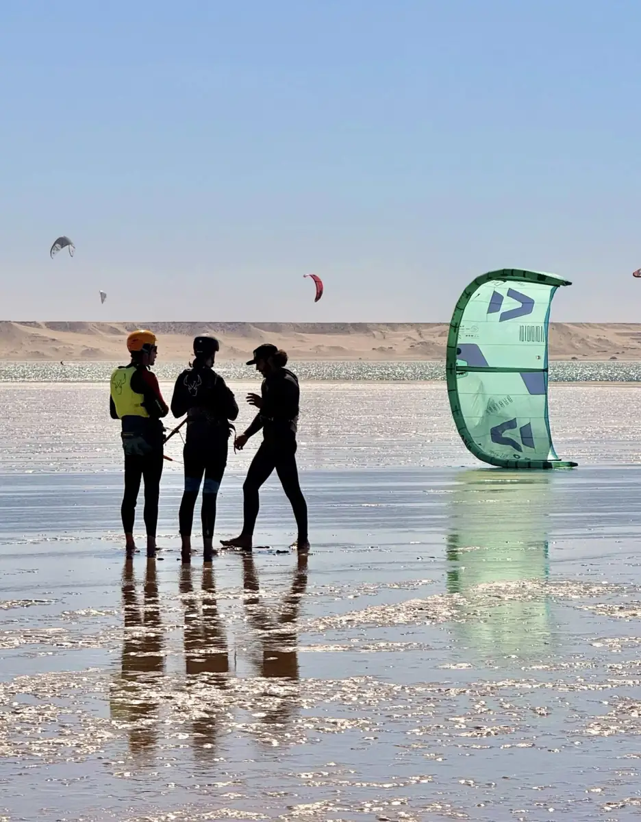 Three people in wetsuits standing on the wet sand beside a kite at the lagoon