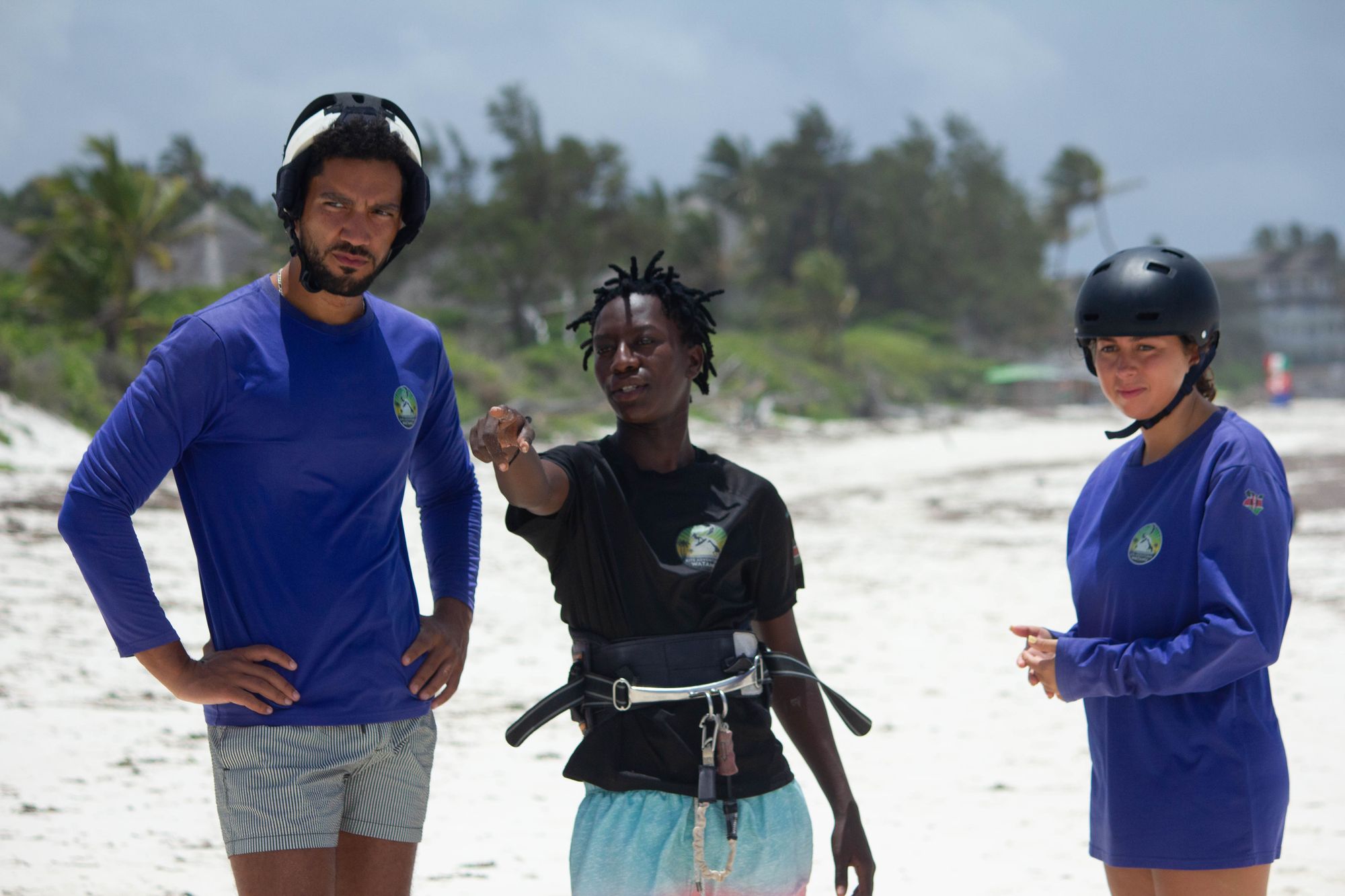 Instructor in a black school shirt explaining something on the beach to two students in helmets