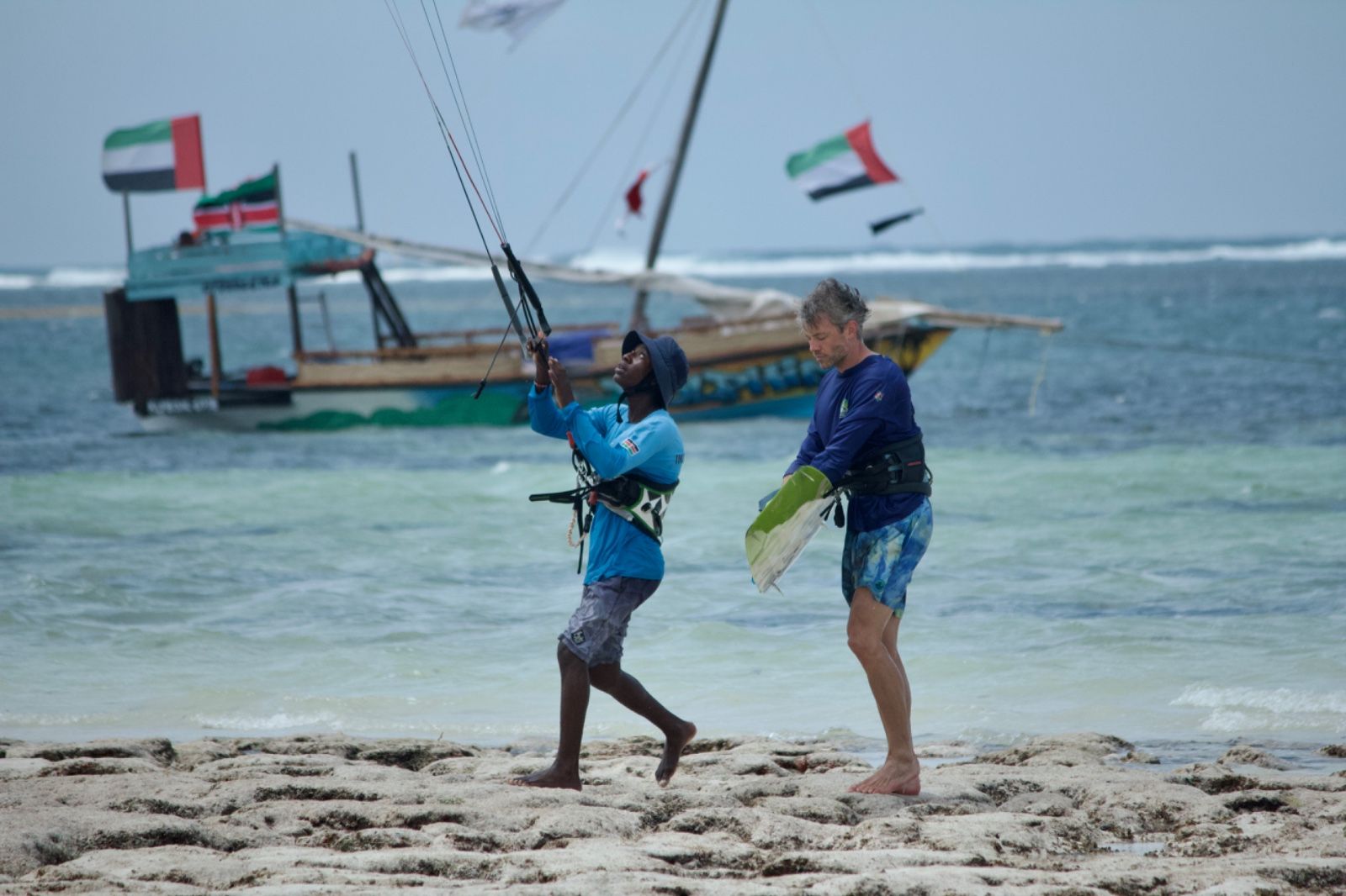 Instructor and student on the beach with a kite, a wooden boat with flags behind them
