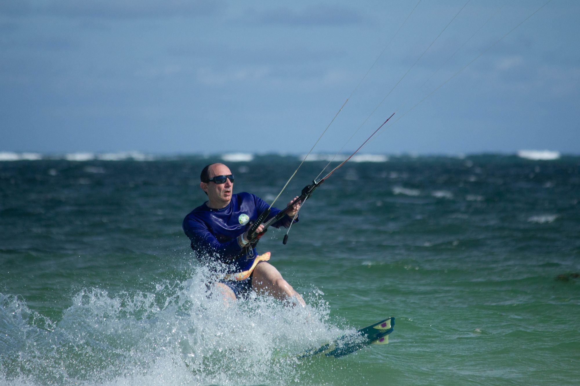 Kitesurfer in a blue shirt and sunglasses riding through white spray