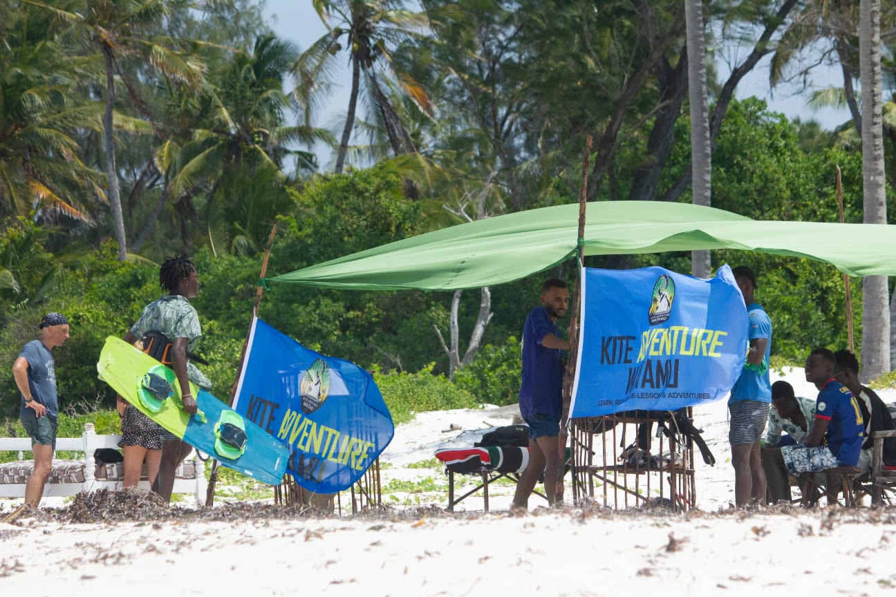 Beach school base with a green canopy, blue Kite Adventure Watamu flags and boards among palm trees