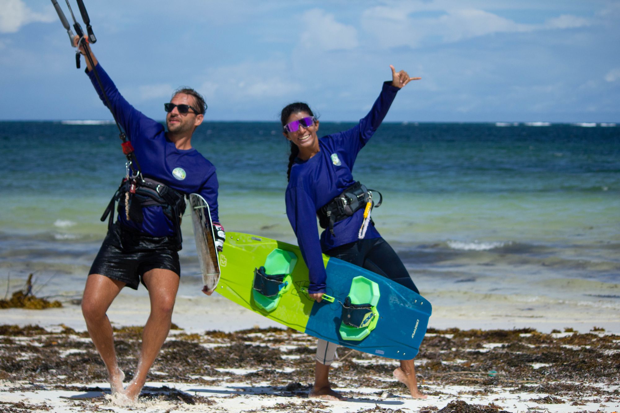 Two kitesurfers in blue school shirts posing on the beach with a board, arms raised