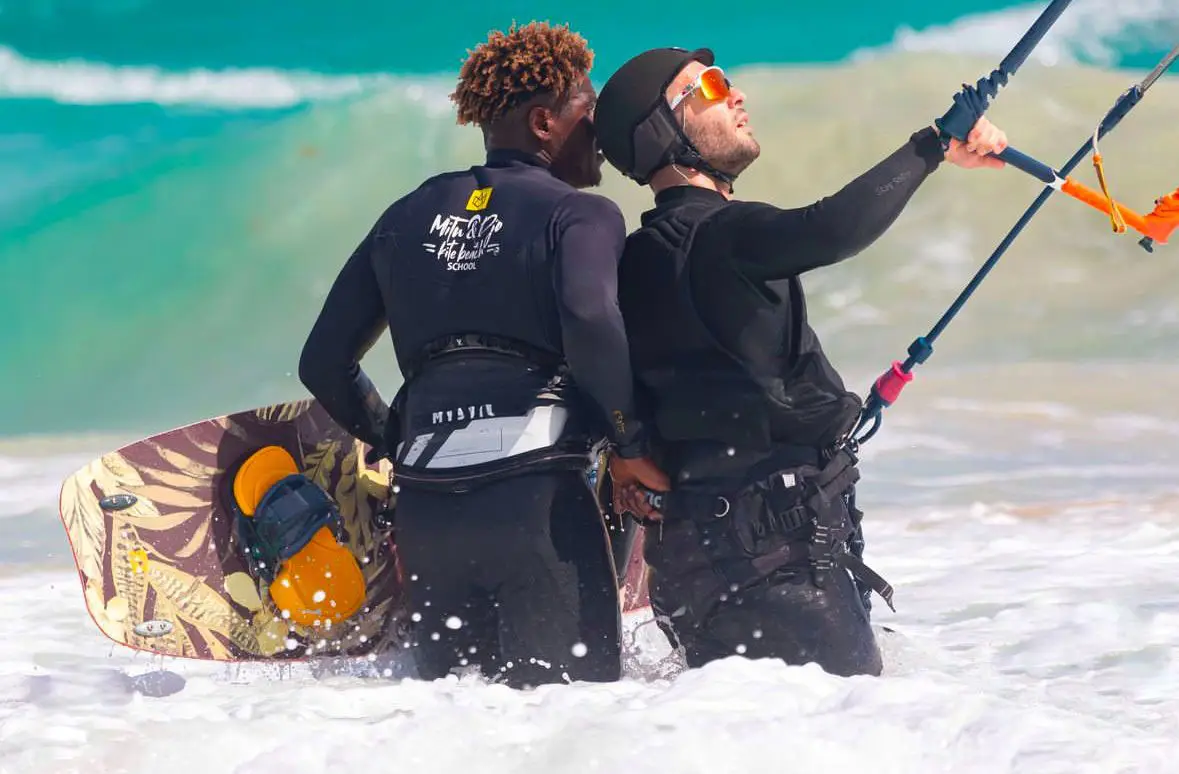 Instructor standing behind a student in a helmet, both holding the control bar in the shore break at Kite Beach