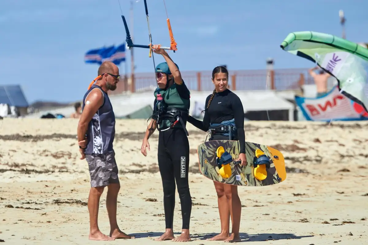 Instructor talking two students through the next step on the beach, one holding the bar and one a board