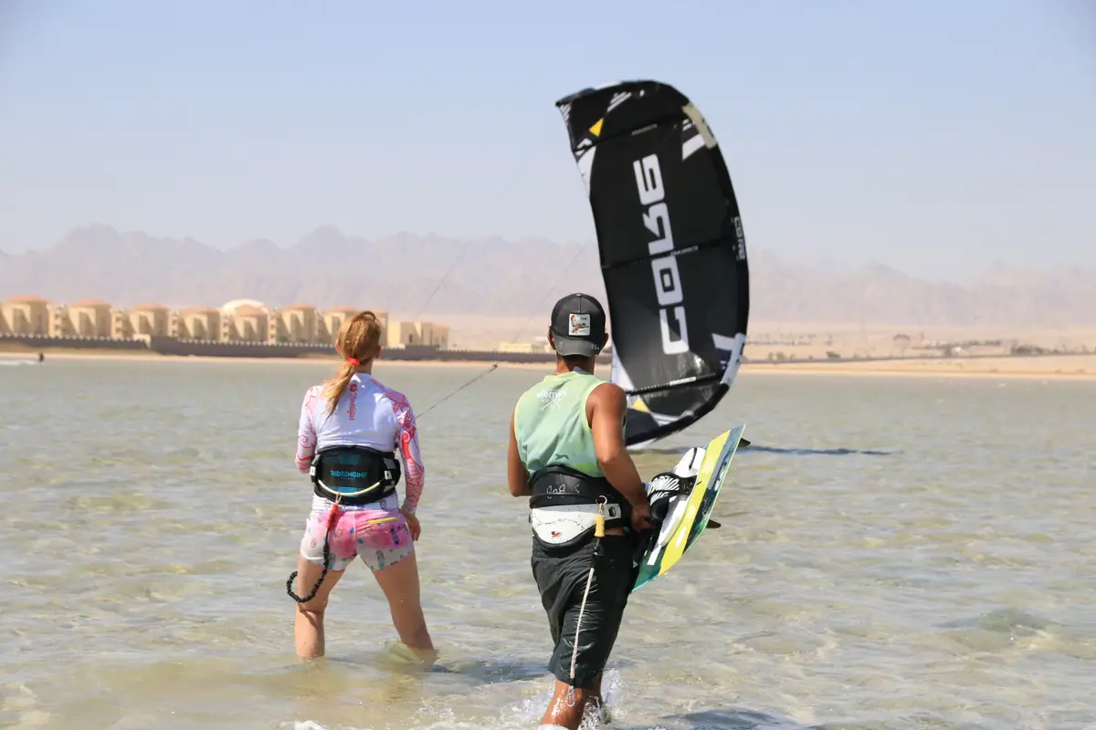 An instructor with a board and a student in a harness wading into the shallow lagoon, a black kite in the air behind them