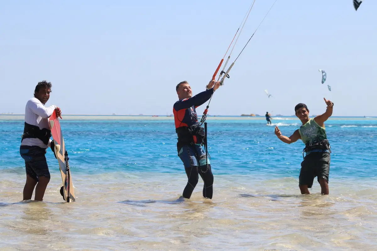 Two instructors and a kitesurf student in the shallow turquoise water, one of them giving a thumbs up, kites in the sky behind
