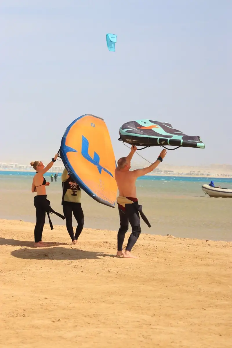 Students and an instructor lifting two wings on the sand at the lagoon, a kite flying in the sky above
