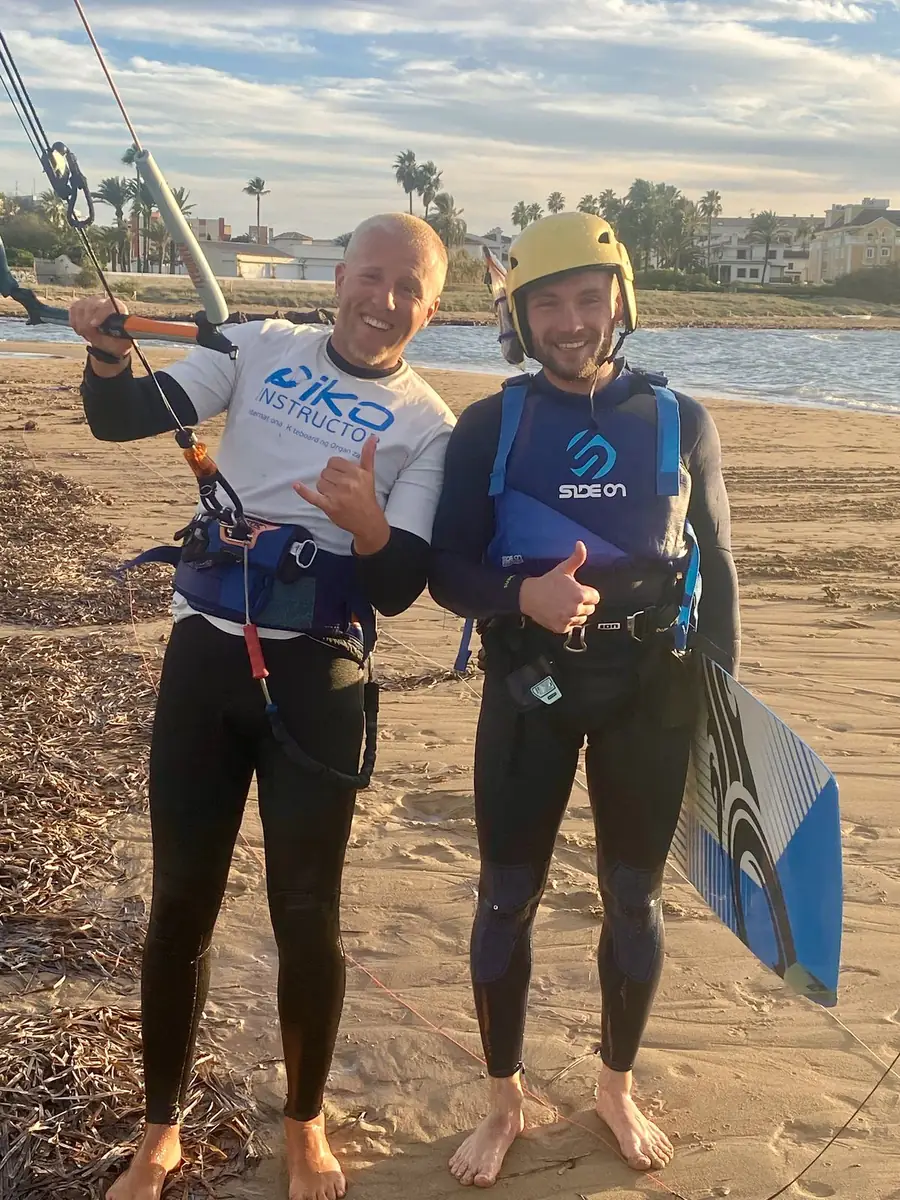Two kitesurfers in IKO instructor and impact vests smiling on the beach at sunset, one holding a kiteboard