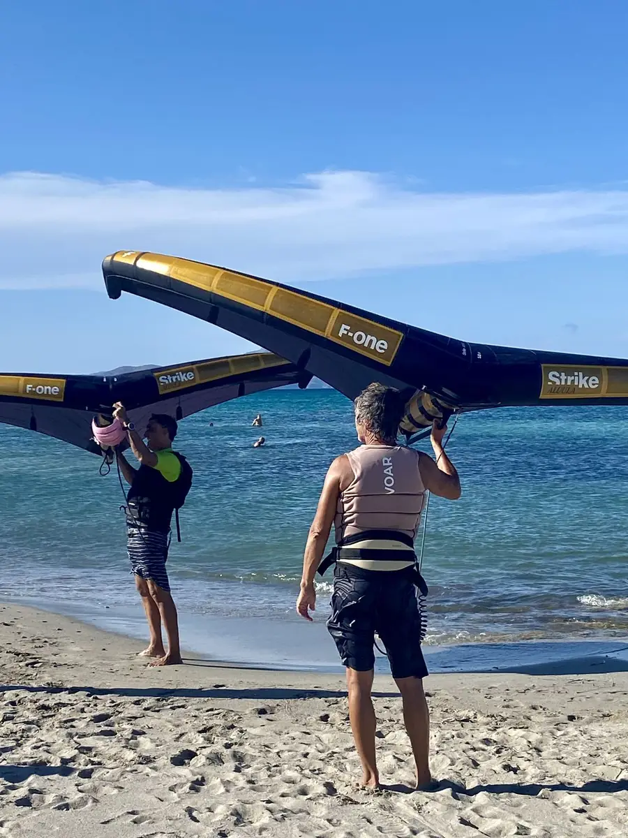 Two people carrying large F-One wings along the shore towards the sea