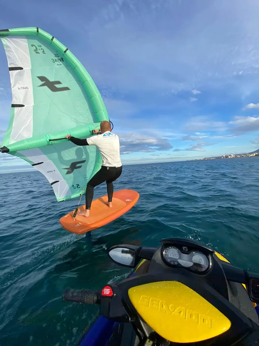 Wingfoiler on an orange board holding a green and white wing, a jet ski in the foreground