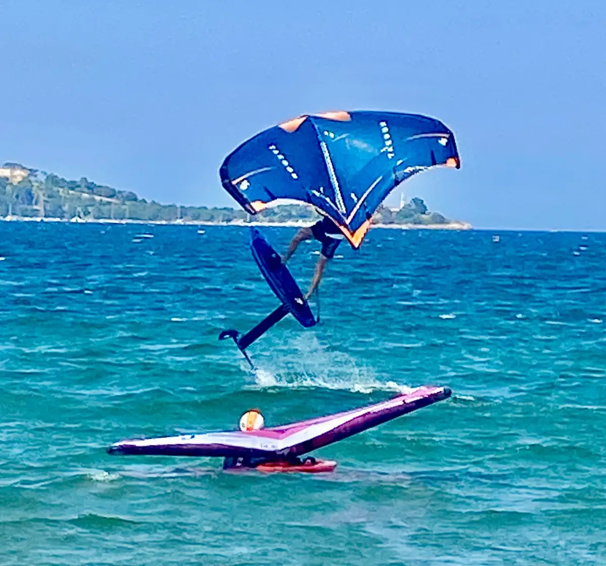 Wingfoiler performing a jump above choppy blue water, a second wing floating on the water below