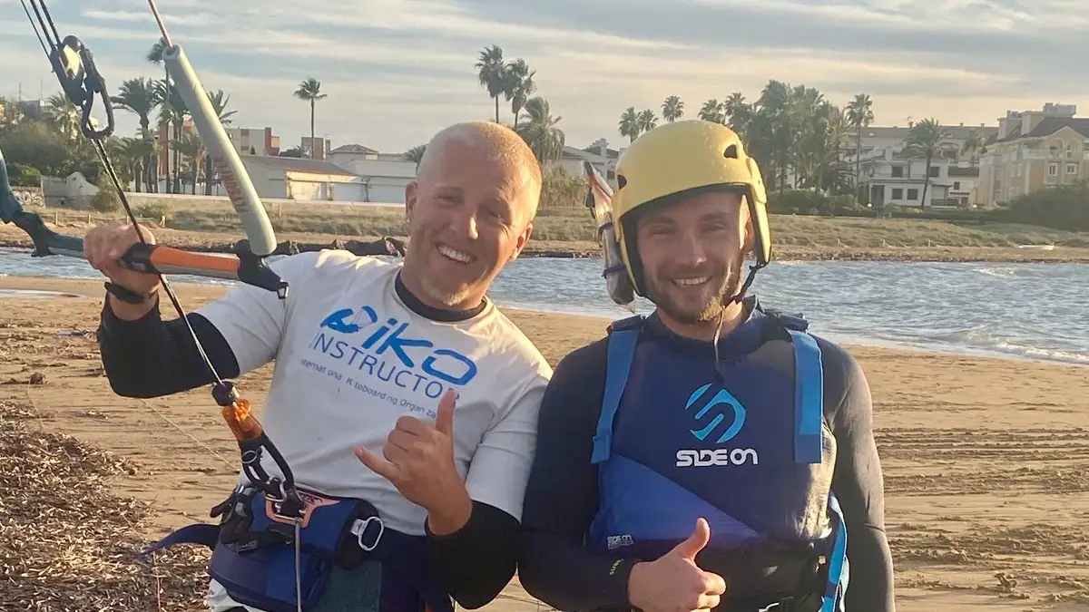 Two smiling kitesurf coaches on the beach, one in an IKO instructor shirt holding the kite bar, one in a yellow helmet, palm trees behind