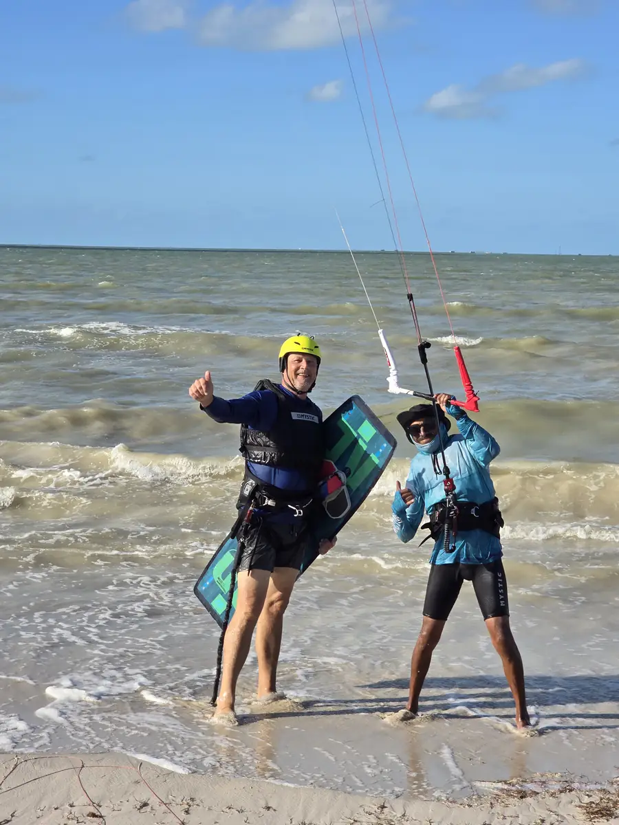 Instructor and a student in a yellow helmet with kite lines and a board, standing in the shallow surf on the beach at Progreso