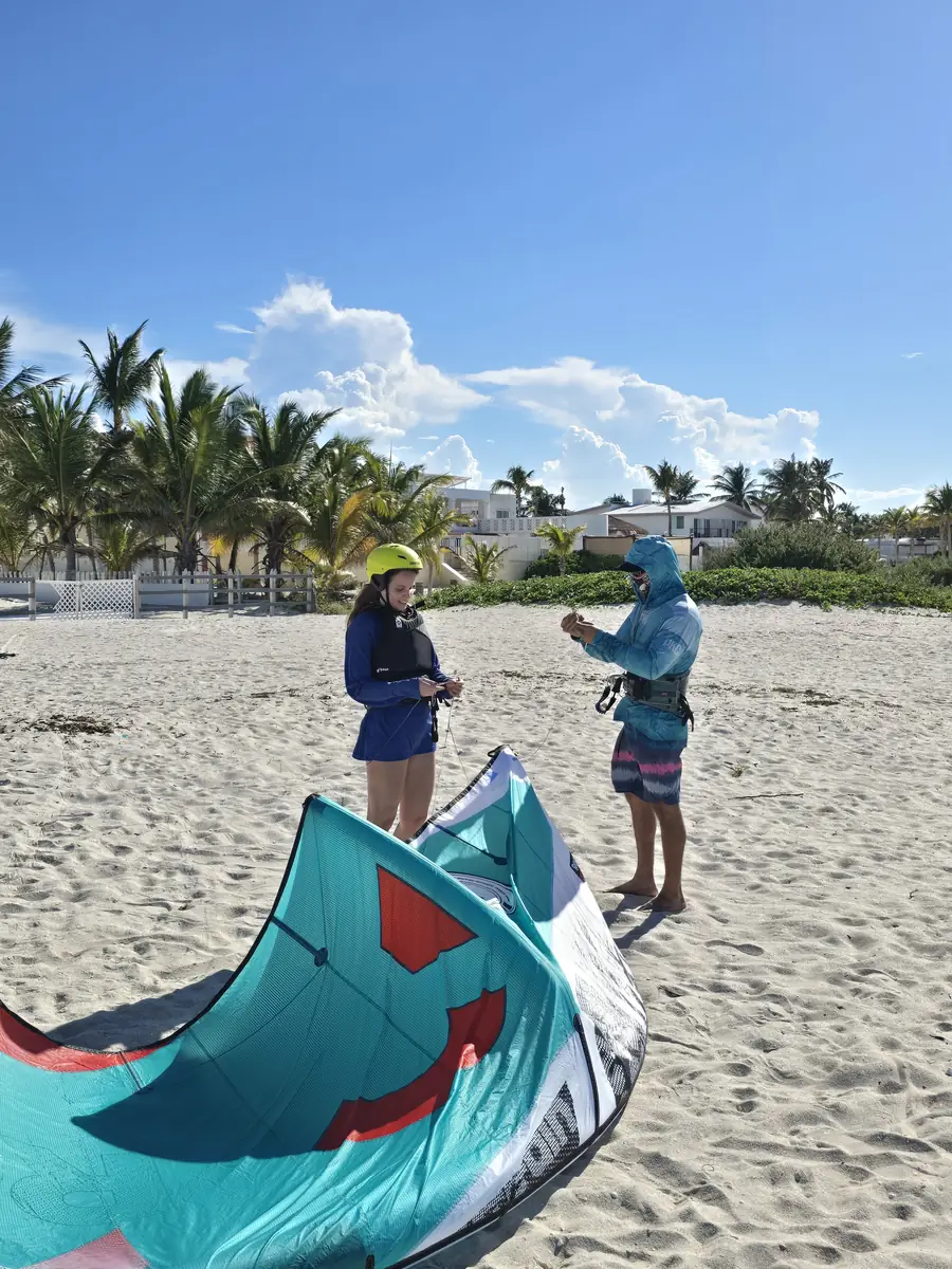Instructor and student with a teal kite spread on the sand, palm trees behind them