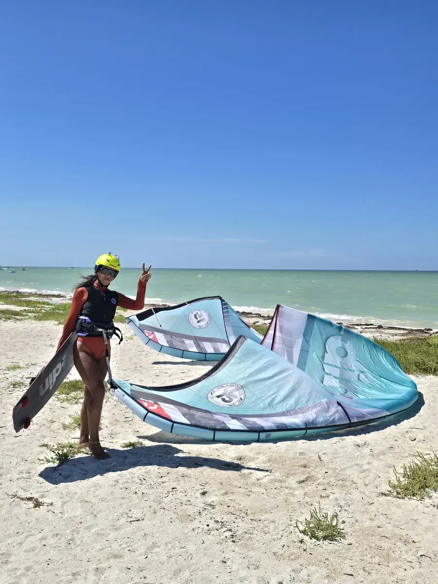 Rider in a yellow helmet holding a board beside two teal kites on the beach