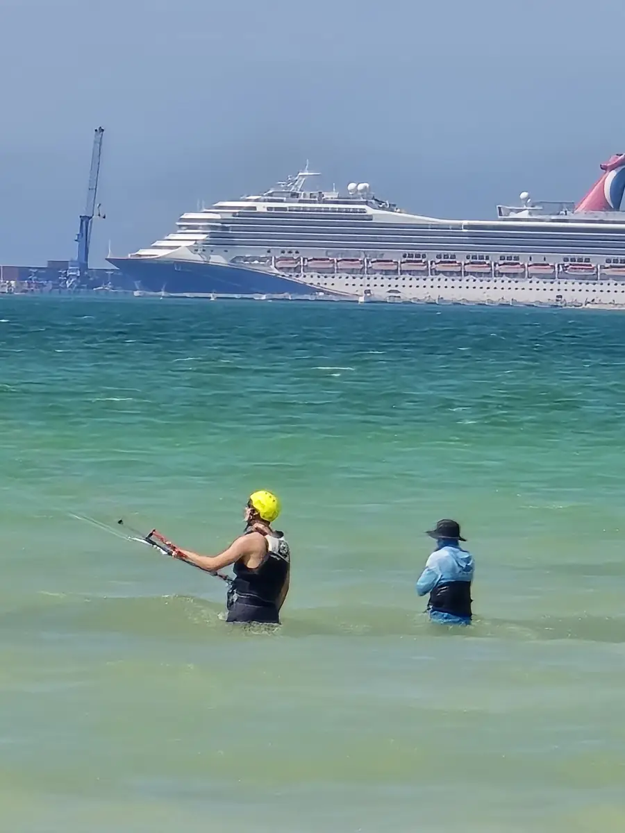 Student in a yellow helmet holding the bar in shallow turquoise water while the instructor watches, a cruise ship in the background