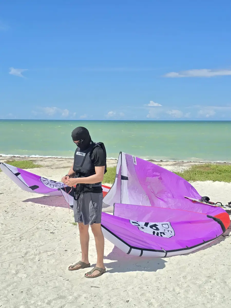 Student in a harness and a face cover next to a purple kite on the white sand, turquoise water behind