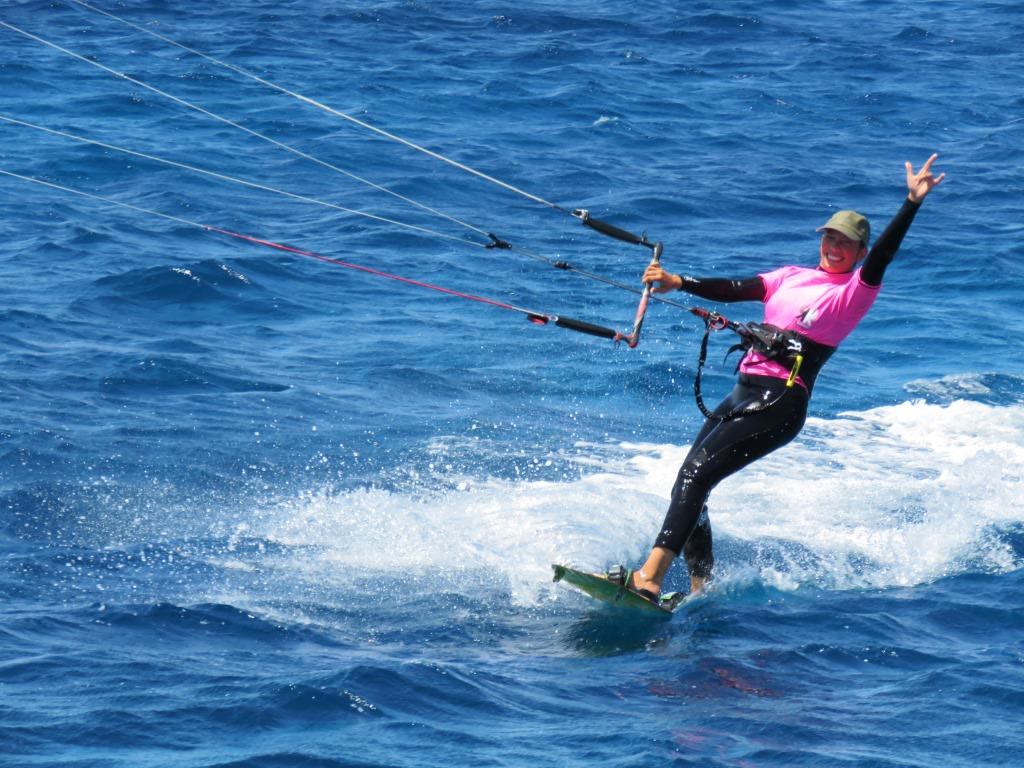 Kitesurfer in a pink top riding fast across deep blue water and waving