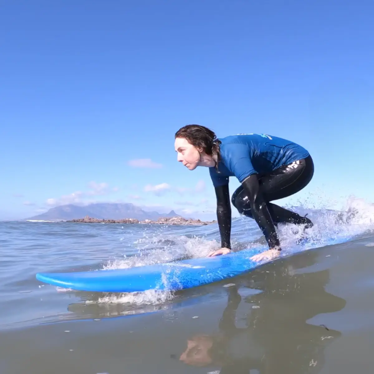 A student in a blue rash vest popping up on a blue board, Table Mountain in the distance