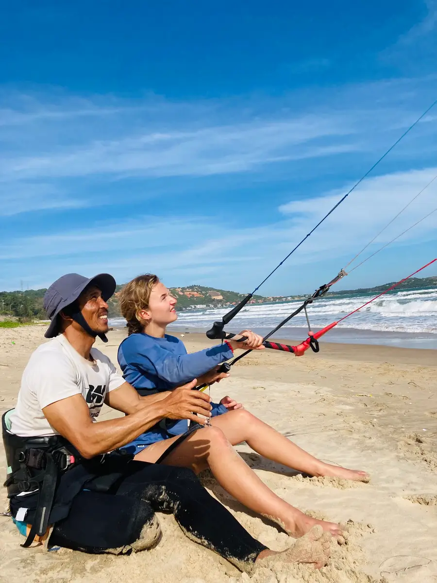 An instructor and a student sitting on the sand in Mui Ne, both holding the kite bar with the lines running up to the kite