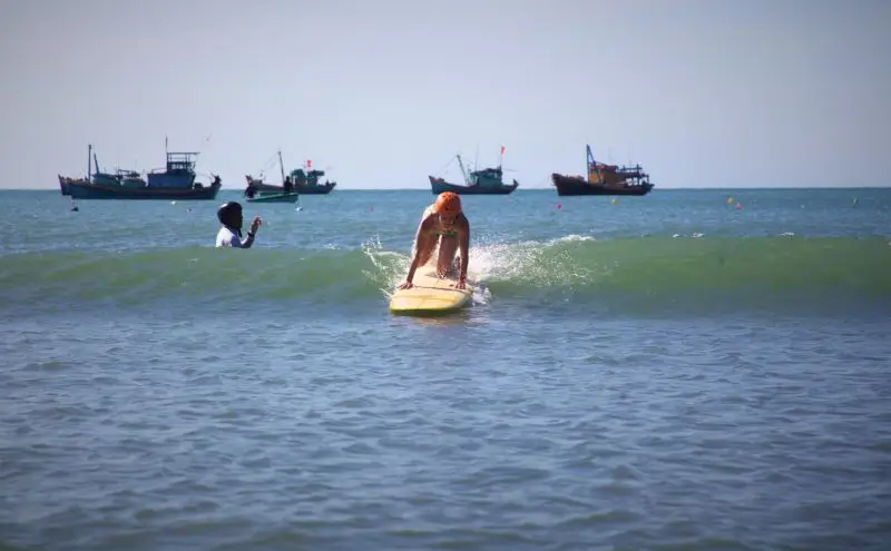 A surfer riding a small wave in Mui Ne with fishing boats on the horizon