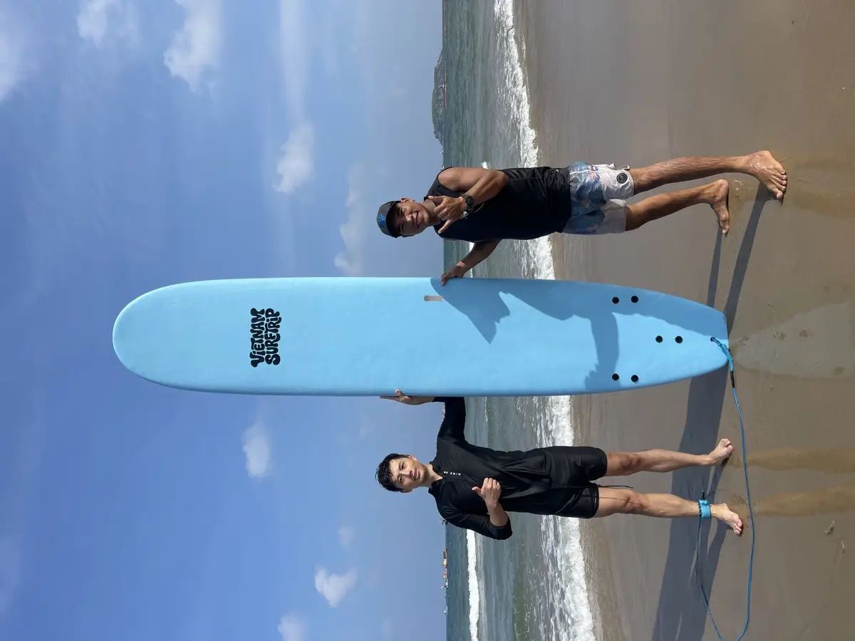 A student and a Vietnam Surftrip coach holding a surfboard upright at the shoreline after a lesson