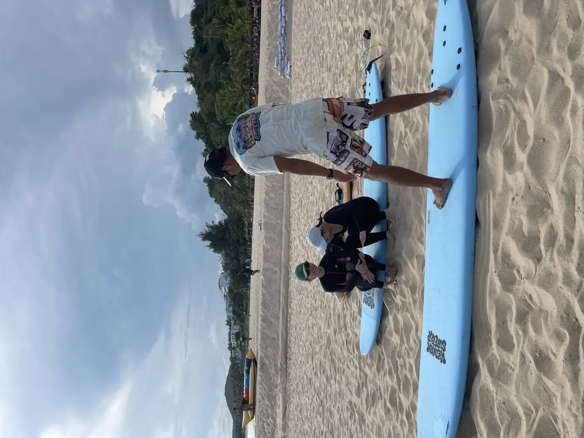 A Vietnam Surftrip coach standing on a soft-top surfboard on the sand to show the pop-up while two students watch